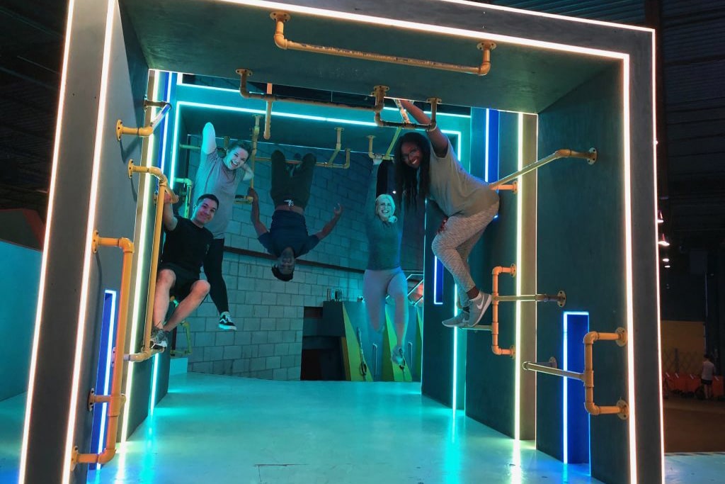 A group of adults enjoying a Ninja Warrior obstacle course in an indoor playground, hanging from colorful pipes and navigating through a vibrant, neon-lit challenge area.