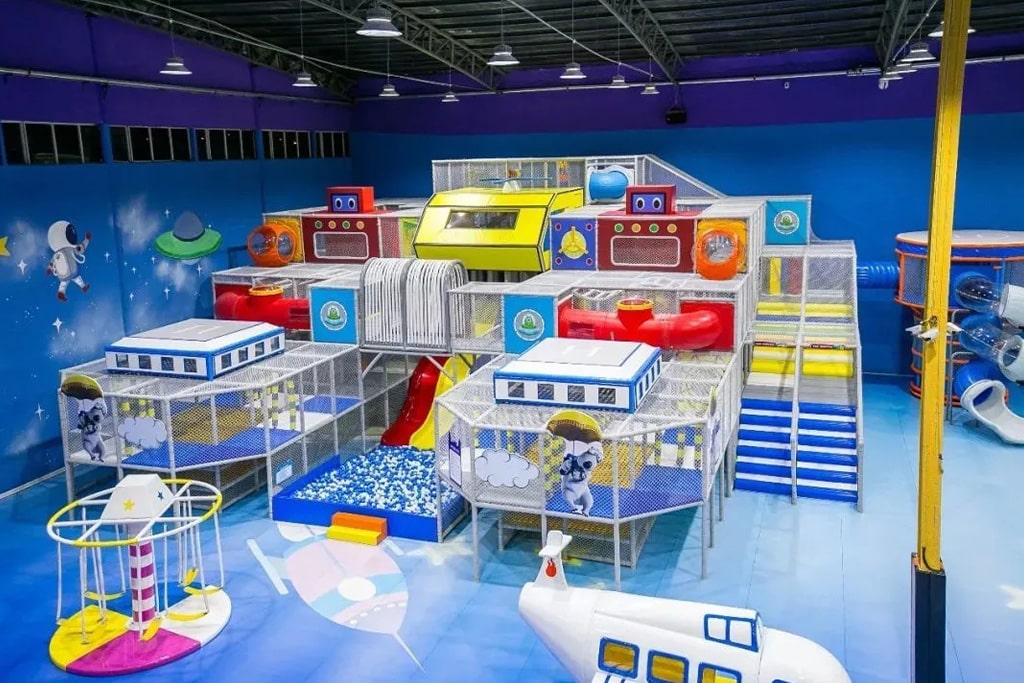 Space-themed indoor playground with rocket-shaped slides, climbing nets, and LED lighting effects.