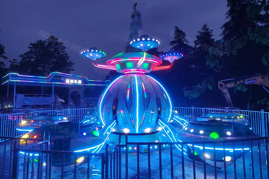 Close-up night photo of Hooplo Space UFO Self Control Plane Ride, colorful LED light interstellar flying saucer kids carnival ride with astronaut statue